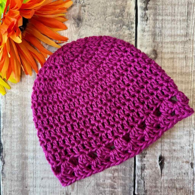 May include: A vibrant magenta beanie crafted with a textured crochet stitch pattern. The hat features a rounded crown and a repeating openwork design along the bottom edge, showcasing a consistent yarn weight and deep, saturated pink color throughout.