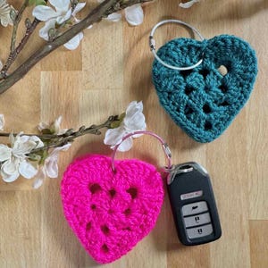 May include: Two handmade crochet heart keychains. One is bright pink, the other is teal. The pink heart is attached to a car key. Both hearts have a textured, open weave design and are attached to silver key rings.