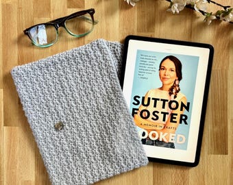 Puede incluir: Una funda de libro de punto gris con un alfiler plateado en la parte delantera. La funda está sobre una superficie de madera junto a una tableta que muestra la portada de un libro titulado "Hooked: A Memoir in Crafts" de Sutton Foster.