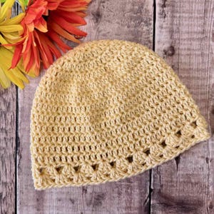 May include: A light yellow crochet beanie hat featuring a textured stitch pattern throughout the main body and a decorative shell-like border along the bottom edge, showcasing a consistent yarn weight and uniform construction.