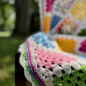 May include: A colorful, handmade crochet blanket draped over a dark wooden chair. The blanket features a patchwork design with squares of pink, green, blue, yellow, and white. The edges are scalloped with a white border and a rainbow stripe.