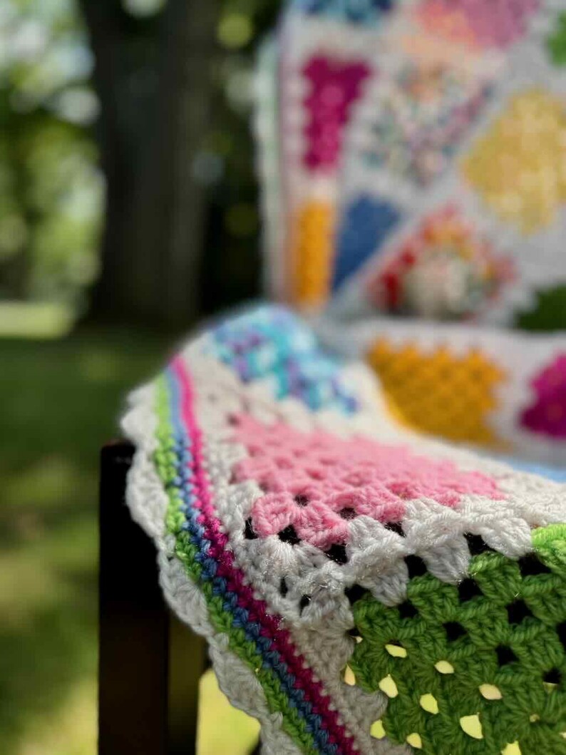 May include: A colorful, handmade crochet blanket draped over a dark wooden chair. The blanket features a patchwork design with squares of pink, green, blue, yellow, and white. The edges are scalloped with a white border and a rainbow stripe.