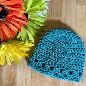 May include: A teal crochet beanie featuring a textured stitch pattern with a repeating row of openwork holes near the bottom edge. The hat is positioned next to a cluster of vibrant orange, yellow, and green artificial flower petals.
