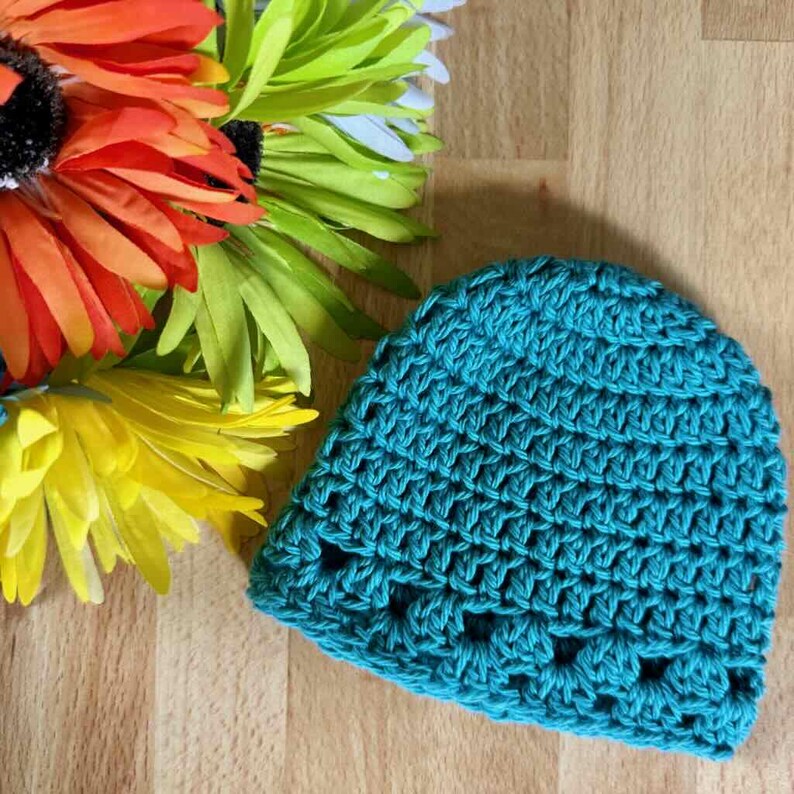 May include: A teal crochet beanie featuring a textured stitch pattern with a repeating row of openwork holes near the bottom edge. The hat is positioned next to a cluster of vibrant orange, yellow, and green artificial flower petals.