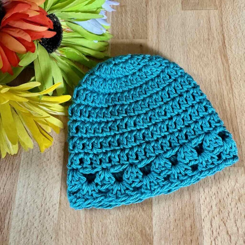 May include: A teal crochet beanie featuring a textured stitch pattern throughout the main body and a decorative shell-like stitch design along the bottom edge. The hat is crafted from yarn and displays a consistent, vibrant blue-green color.
