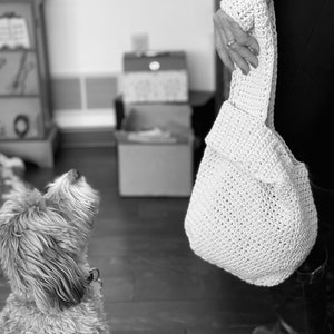 May include: A white crocheted tote bag with a slouchy design. The bag is being held by a person's hand.