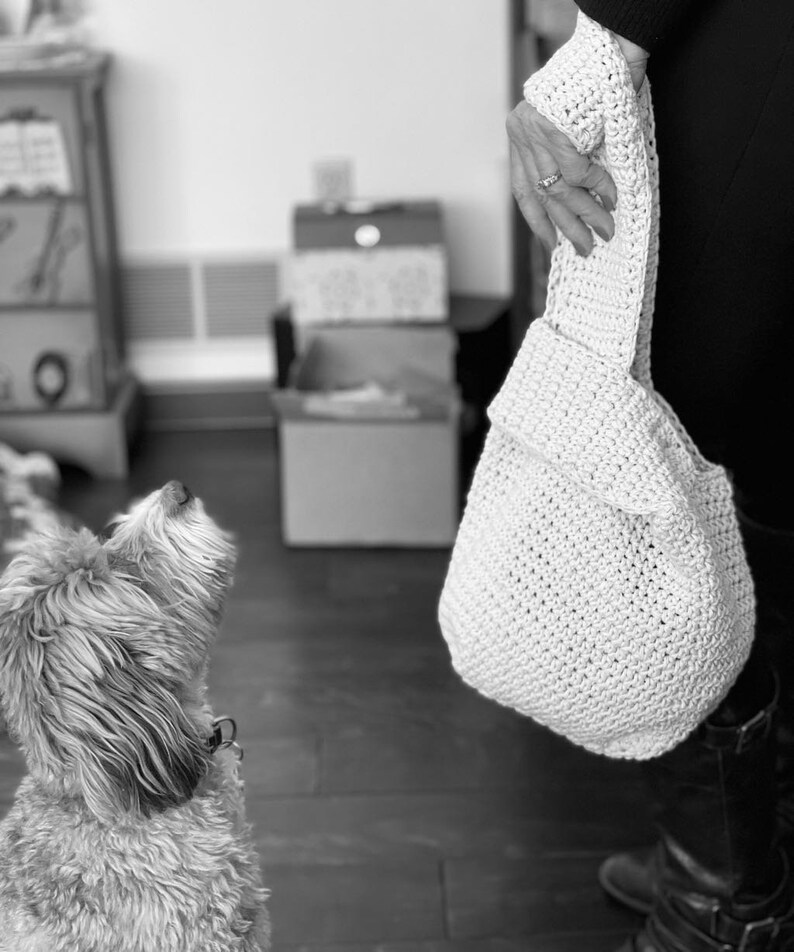 May include: A white crocheted tote bag with a slouchy design. The bag is being held by a person's hand.