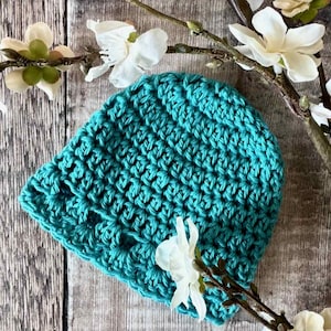 May include: A teal crochet beanie hat featuring a textured, repeating stitch pattern. The hat is crafted from yarn and displays a consistent, dense weave throughout its rounded shape, providing a solid color appearance in a vibrant shade of blue-green.