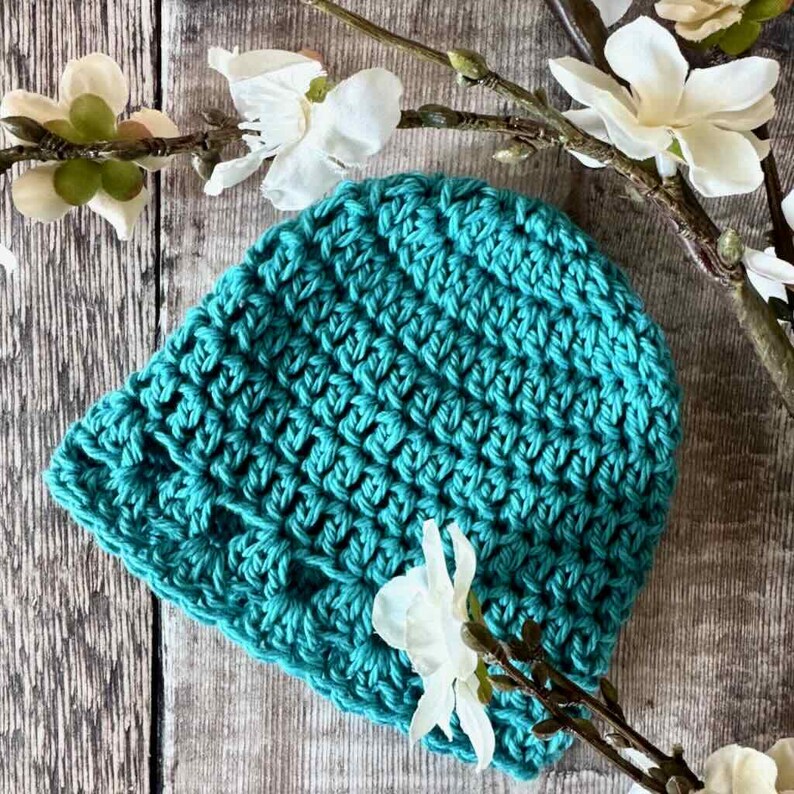 May include: A teal crochet beanie hat featuring a textured, repeating stitch pattern. The hat is crafted from yarn and displays a consistent, dense weave throughout its rounded shape, providing a solid color appearance in a vibrant shade of blue-green.