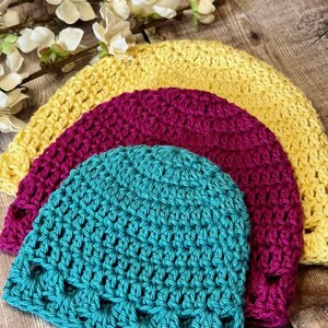 May include: Three crocheted beanies stacked together, featuring a bright yellow hat on the bottom, a deep magenta hat in the middle, and a vibrant turquoise hat on top. Each hat is crafted with a consistent, textured stitch pattern.