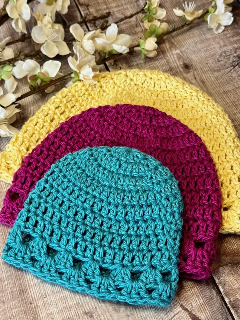 May include: Three crocheted beanies stacked together, featuring a bright yellow hat on the bottom, a deep magenta hat in the middle, and a vibrant turquoise hat on top. Each hat is crafted with a consistent, textured stitch pattern.