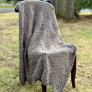May include: A brown crocheted throw blanket with a textured pattern is draped over a wooden chair. The chair is sitting on a grassy lawn.