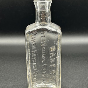 May include: A clear, rectangular glass bottle with a narrow neck. The bottle has embossed text that reads "BAKER'S FLAVORING EXTRACT COMPANY". The bottle is empty and appears to be an antique.