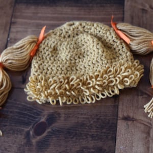 May include: A crocheted hat with two long blonde yarn pigtails tied with orange ribbons. The hat has a ruffled edge.