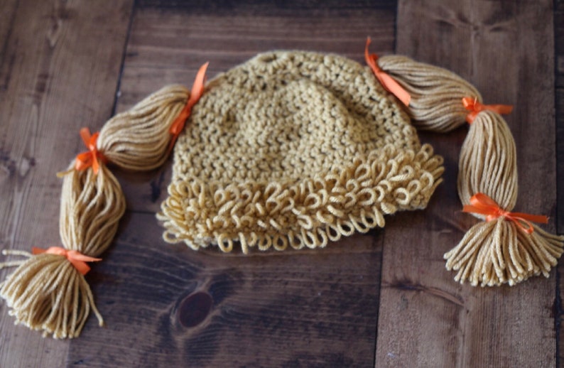 May include: A crocheted hat with two long blonde yarn pigtails tied with orange ribbons. The hat has a ruffled edge.