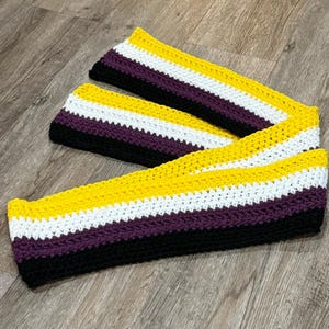 May include: A crocheted scarf with yellow, white, purple, and black stripes. The scarf is folded in half and the ends are overlapping.