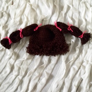 May include: Brown crocheted doll hat with two brown yarn pigtails tied with pink ribbons.