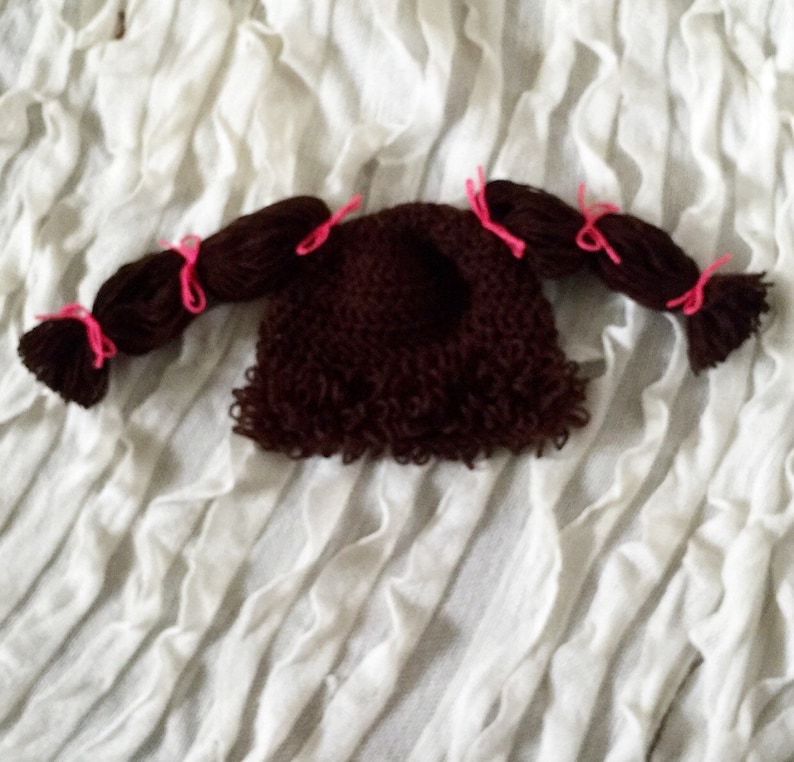 May include: Brown crocheted doll hat with two brown yarn pigtails tied with pink ribbons.