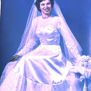 May include: A bride wearing a white satin wedding dress with a lace trim and a veil. She is holding a bouquet of white flowers.