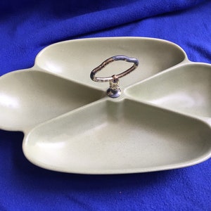 May include: A green ceramic serving dish with four sections and a gold metal handle in the center. The dish is shaped like a four-leaf clover.