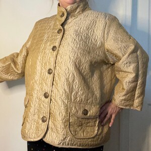May include: A quilted jacket in a light gold color with a stand-up collar and a button closure. The jacket has a patch pocket on the left side and a decorative pattern throughout.