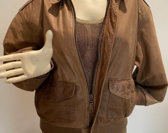 May include: A brown leather bomber jacket with a zip-up front, two front pockets, and a ribbed hem and cuffs. The jacket is displayed on a mannequin. The jacket has a collared neckline and shoulder epaulets.