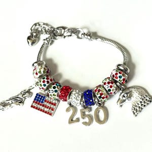 May include: A silver-tone charm bracelet featuring a series of beads encrusted with red, white, and blue rhinestones. The bracelet includes a rectangular American flag charm, a silver eagle head charm, a Statue of Liberty charm, and the number 250.