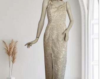 May include: A gold-colored, floor-length dress with a floral pattern and a front slit. The dress has thin straps and a sweetheart neckline. The dress is displayed on a mannequin in front of a white background.