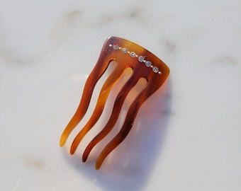 May include: A tortoiseshell hair comb with four curved tines. The comb is decorated with a row of small, clear rhinestones along the top edge. The comb is a warm brown color with lighter amber highlights.