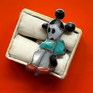 May include: A silver-toned pin in the shape of a stylized figure, featuring a white face, black ears, and turquoise and red accents. The pin is set against a white background inside a small box, with an orange background.