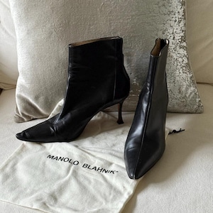 Puede incluir: Botines de cuero negro con punta afilada y tacones de aguja. Las botas están sobre una superficie crema. Una bolsa de tela blanca con el texto "MANOLO BLAHNIK" está en la parte inferior. Los tacones miden unos 8 cm.