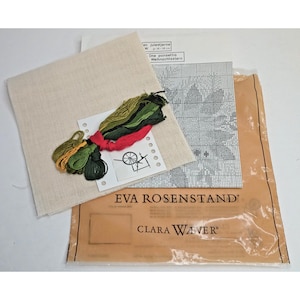 May include: A cross-stitch kit featuring a beige fabric, a pattern, and colorful embroidery floss in shades of green and red. The kit includes a paper with the text "EVA ROSENSTAND" and "CLARA WAVER".
