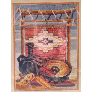 May include: A framed needlepoint artwork depicting a Southwestern scene. The artwork features a red and white patterned rug, a black vase, and a bowl with a colorful design. The background includes a wooden structure and a blue sky.