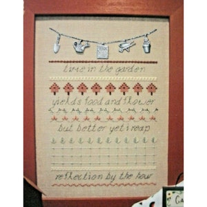 May include: A framed cross-stitch sampler with the text "Time in the garden yields food and flower but better yet I reap reflection by the hour". The sampler features a chain of charms with gardening tools and a flower pot.
