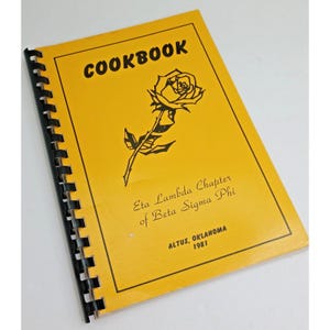 May include: A yellow spiral-bound cookbook with black text and a rose illustration. The cover reads COOKBOOK, Eta Lambda Chapter of Beta Sigma Phi, and ALTUS, OKLAHOMA 1981. The book features a black plastic comb binding along the left side edge.