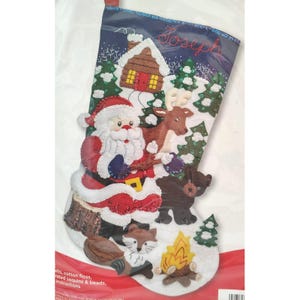 May include: A felt Christmas stocking kit featuring the name Joseph embroidered in red at the top. The design includes a Santa Claus figure, a brown reindeer, a black bear, a fox, a log cabin, and a campfire, all decorated with sequins and beads.