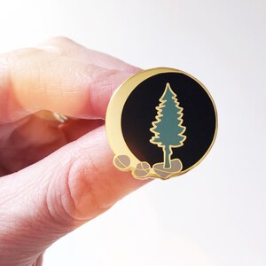 May include: Gold-tone enamel pin featuring a green pine tree silhouette on a black background with a crescent moon shape. The tree is standing on three small brown rocks.
