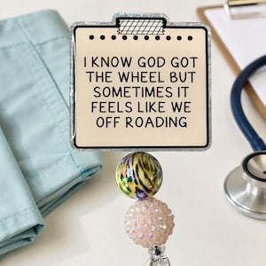 May include: A square badge reel featuring a cream-colored face with the black text I KNOW GOD GOT THE WHEEL BUT SOMETIMES IT FEELS LIKE WE OFF ROADING. Below the square are two decorative beads, one multicolored and one pale pink, above a clear clip.