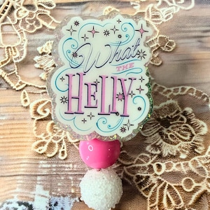 May include: A badge reel with a clear, glittery top featuring the phrase "What the Helly" in a whimsical font. The reel includes a pink heart, a white pom-pom, and a glittery, round clip.