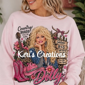 May include: A light pink sweatshirt featuring a graphic print of a blonde woman, a guitar, pink cowboy boots, a pink hat, and a cow skull. The text includes Country Roads Take Me Home, Jolene, 9 to 5, I Will Always Love You, Dolly, and Keri's Creations.