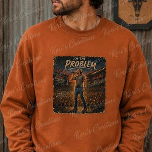 Pode incluir: Uma camisola de gola redonda em cor laranja queimado com uma impressão gráfica de uma cena de concerto com um artista no palco. O texto "I'M THE PROBLEM" está impresso acima da ilustração, que mostra uma multidão num estádio.