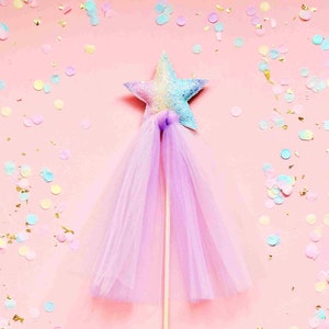 May include: A pink and purple star-shaped wand with a tulle skirt and a wooden stick. The wand is surrounded by colorful confetti on a pink background.