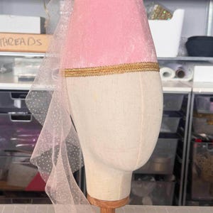 A pink velvet hat with a gold trim is displayed on a mannequin head in a workshop setting.