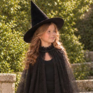 May include: A black pointed witch hat made of velvet material paired with a sheer black cape featuring a delicate dotted pattern and a ruffled collar. The cape is worn over a simple black top, creating a cohesive dark ensemble for costume wear.