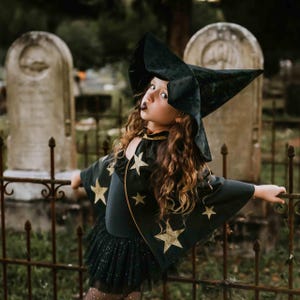 May include: A child in a black witch costume with a large, pointed hat and a cape adorned with gold stars. The costume includes a black tutu skirt. The background features a wrought iron fence and weathered tombstones, creating a Halloween theme.