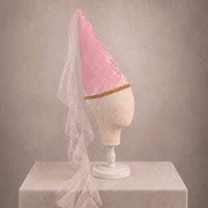 A pink velvet hat with a sheer veil draped over the front, displayed on a white mannequin head stand against a neutral background.
