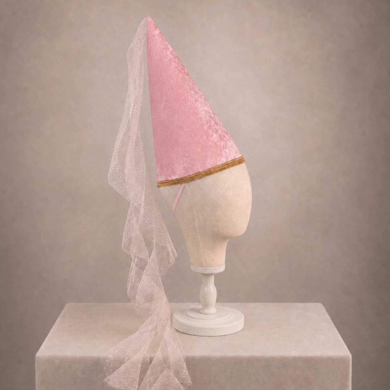 A pink velvet hat with a sheer veil draped over the front, displayed on a white mannequin head stand against a neutral background.