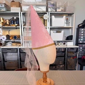 A pink velvet hat with a gold trim is displayed on a wooden stand in a cluttered room filled with various items and storage containers.