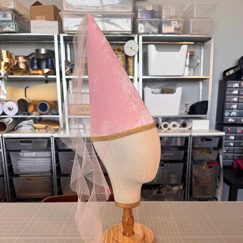 A pink velvet hat with a gold trim is displayed on a wooden stand in a cluttered room filled with various items and storage containers.