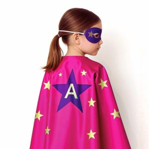 May include: A pink superhero cape with a large purple star featuring the letter "A" and gold star accents. A matching purple eye mask with gold star details completes the costume. The cape is made of a shiny material.